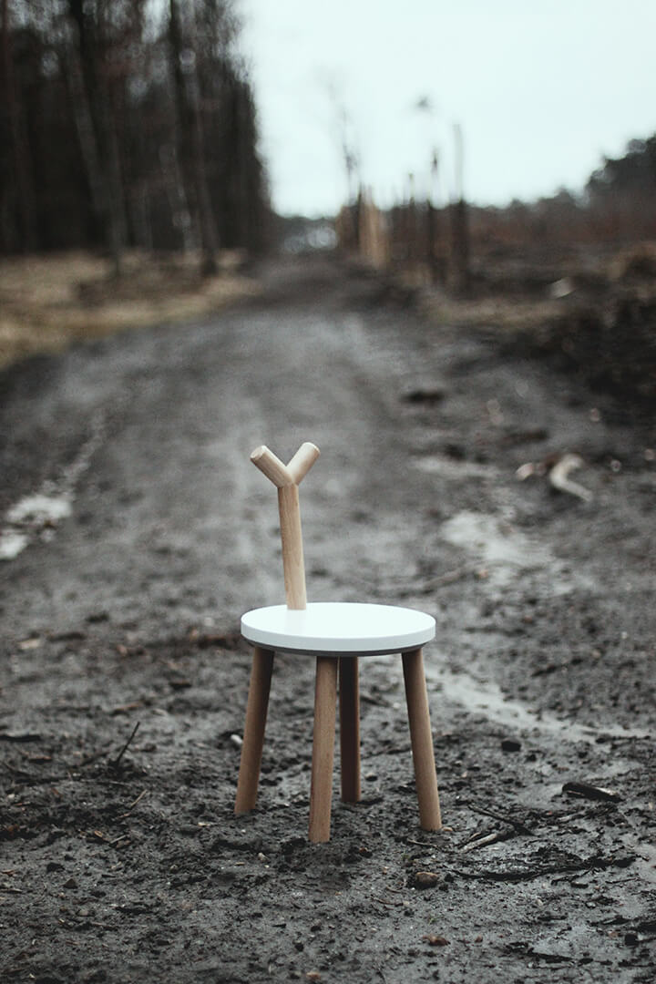 dziecięcy drewniany stołek z czterema nogami, białym siedziskiem i oparciem przypominającym główkę jelonka stoi na błotnistej drodze w lesie
