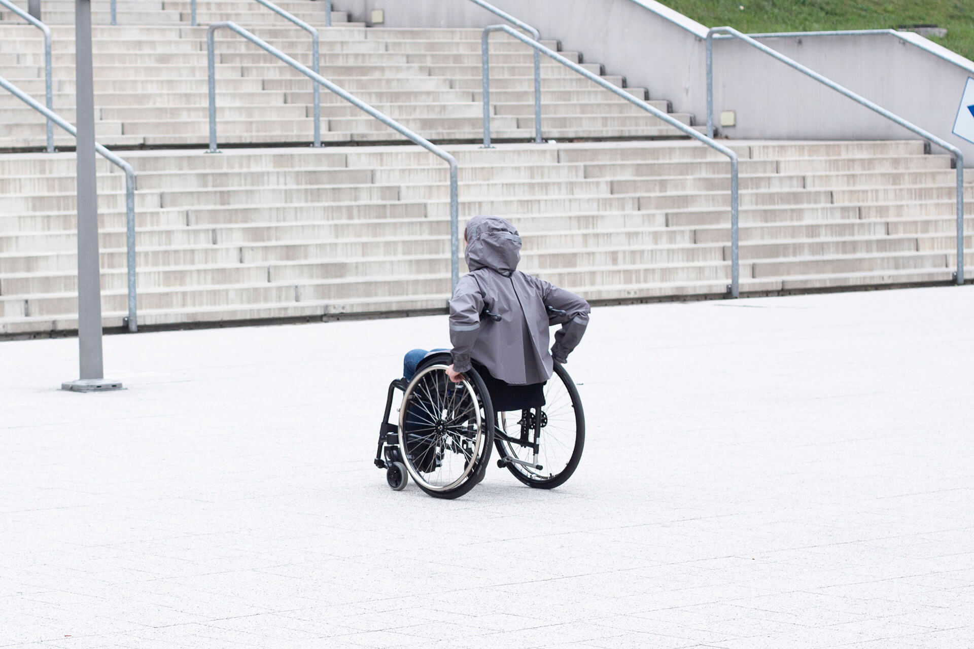 osoba na wózku inwalidzkim ubrana w płaszcz przeciwdeszczowy, który jest zaprojektowany tak, że nie krępuje jej ruchów, w tle nowoczesna architektura, betonowe schody