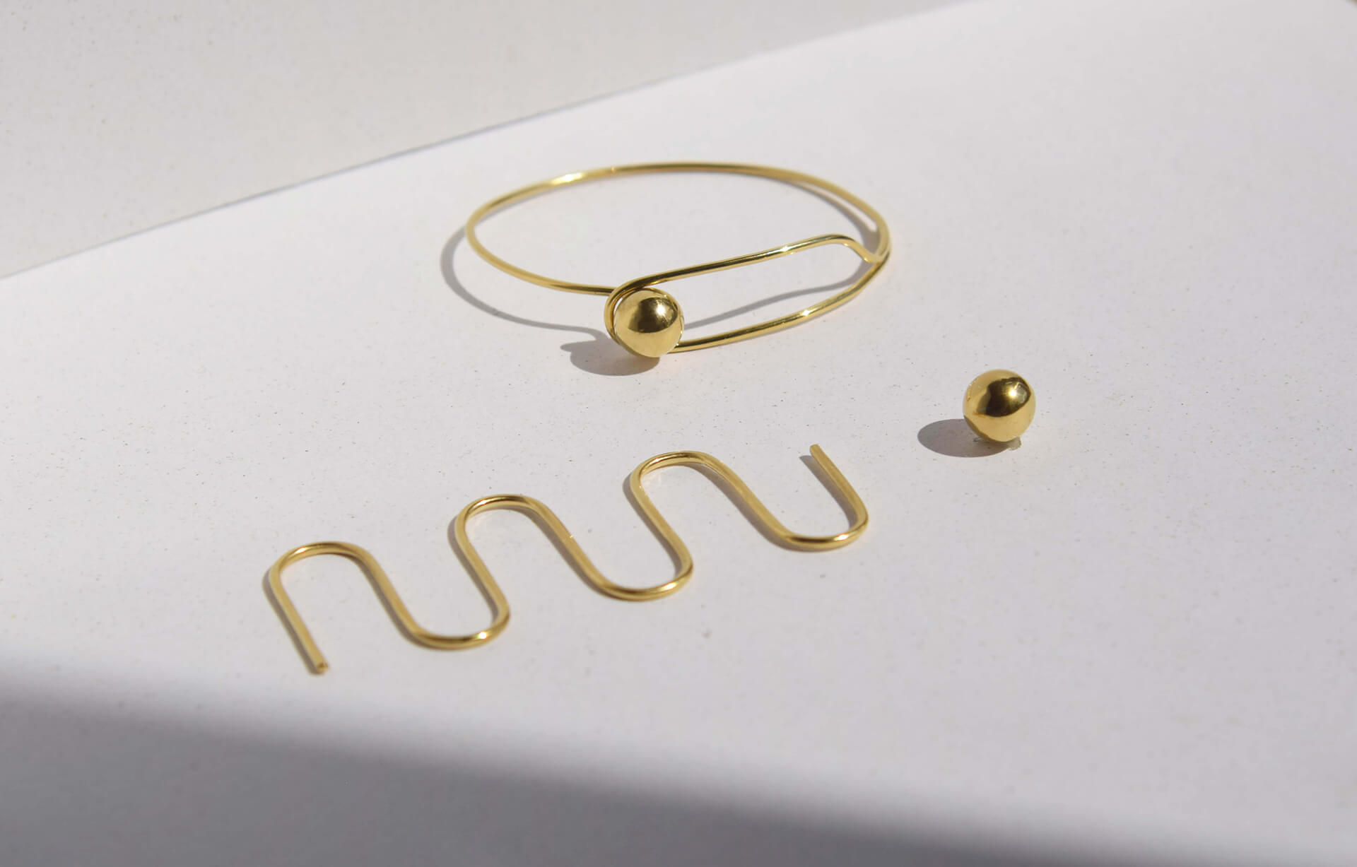 Złota biżuteria: z element o kształcie wijącej się linii, za nim złota kulka, z tyłu pierścionek z kulą otoczoną pętlą